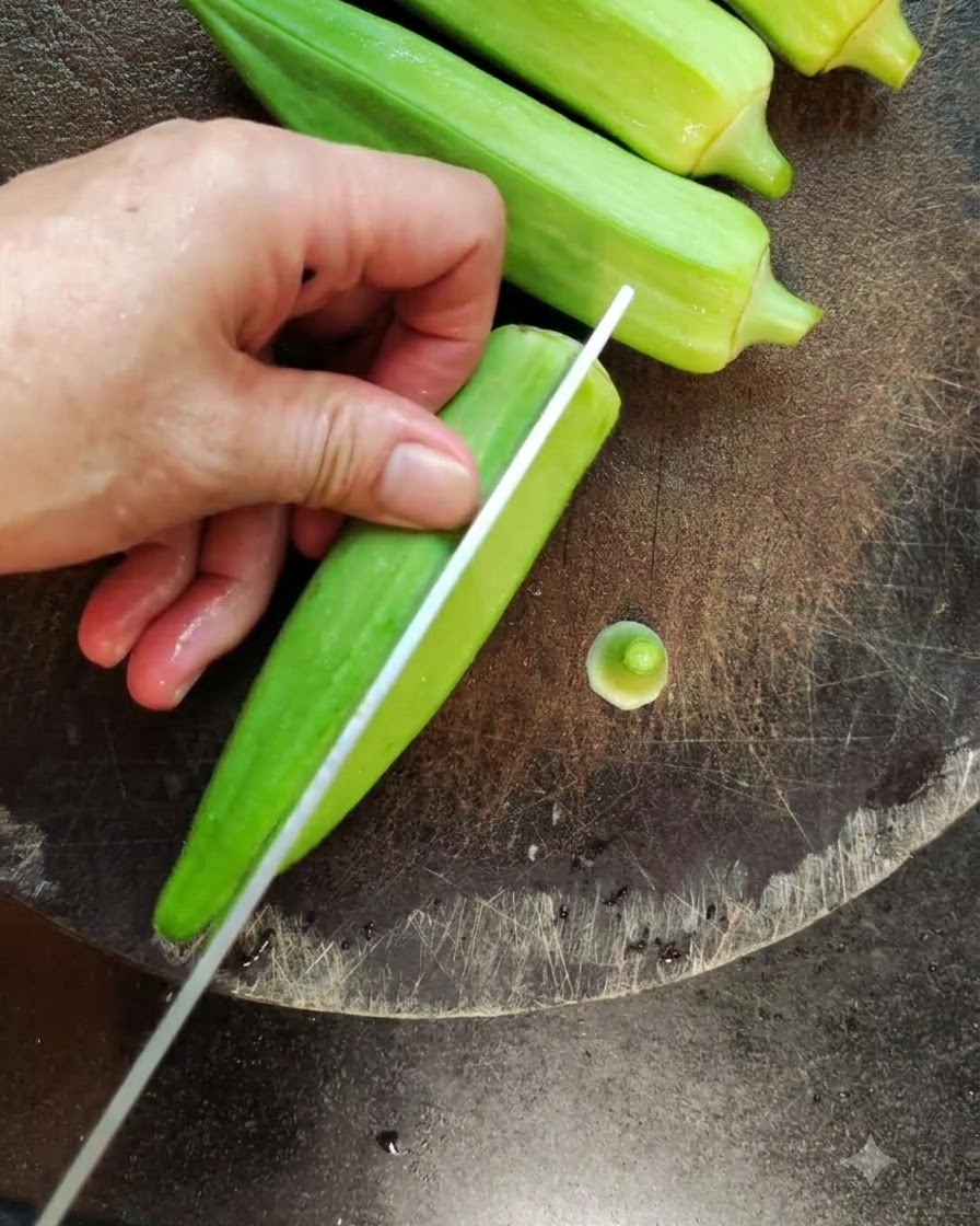 Eine Nahaufnahme eines Messers, das eine frische grüne Okra der Länge nach auf einem dunklen Schneidebrett teilt.
