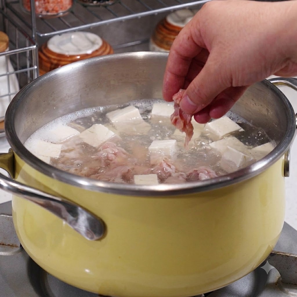 Eine Hand legt marinierte Schweinefleischscheiben in einen Topf mit kochender Brühe, Tofu und Meeresfrüchten.
