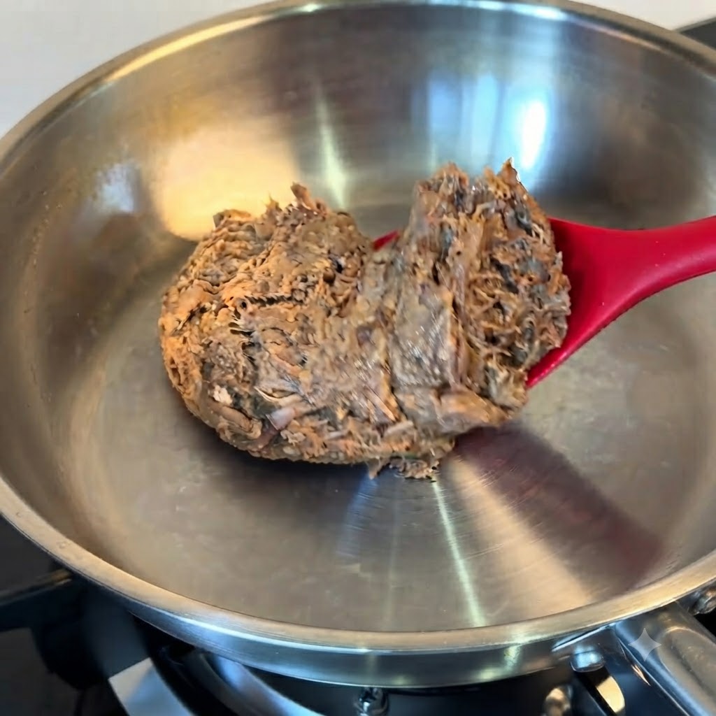 Ein Block aufgetautes Pulled Beef wird mit einem roten Spatel in eine Edelstahlpfanne gegeben.