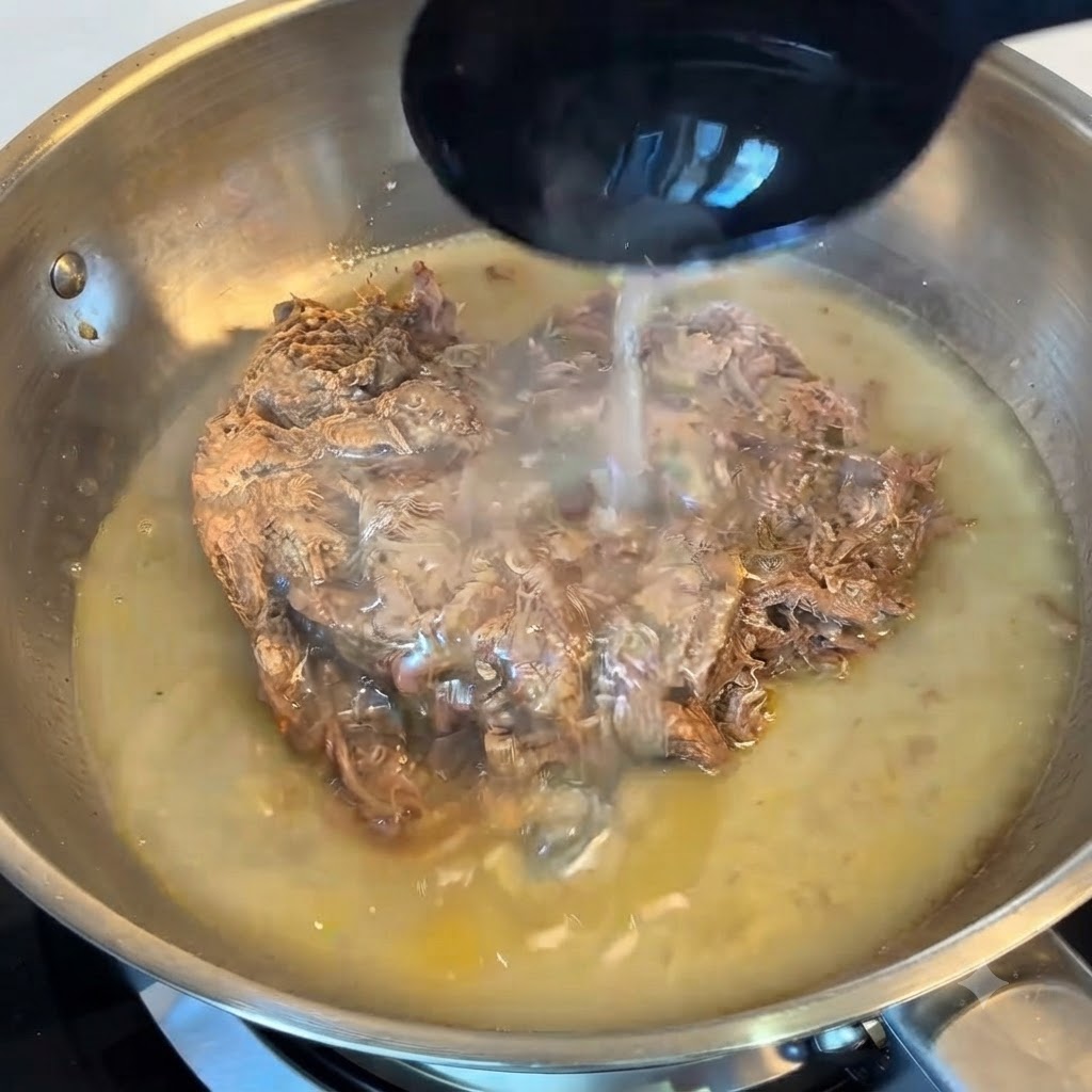 Eine Schöpfkelle gießt stärkehaltiges Nudelwasser über das Pulled Beef in einer Edelstahlpfanne.