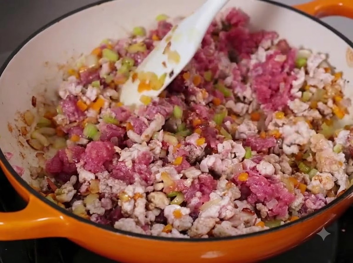 Rinder- und Schweinehackfleisch wird mit gewürfeltem Gemüse in einer großen Pfanne mit einem weißen Spatel vermischt.