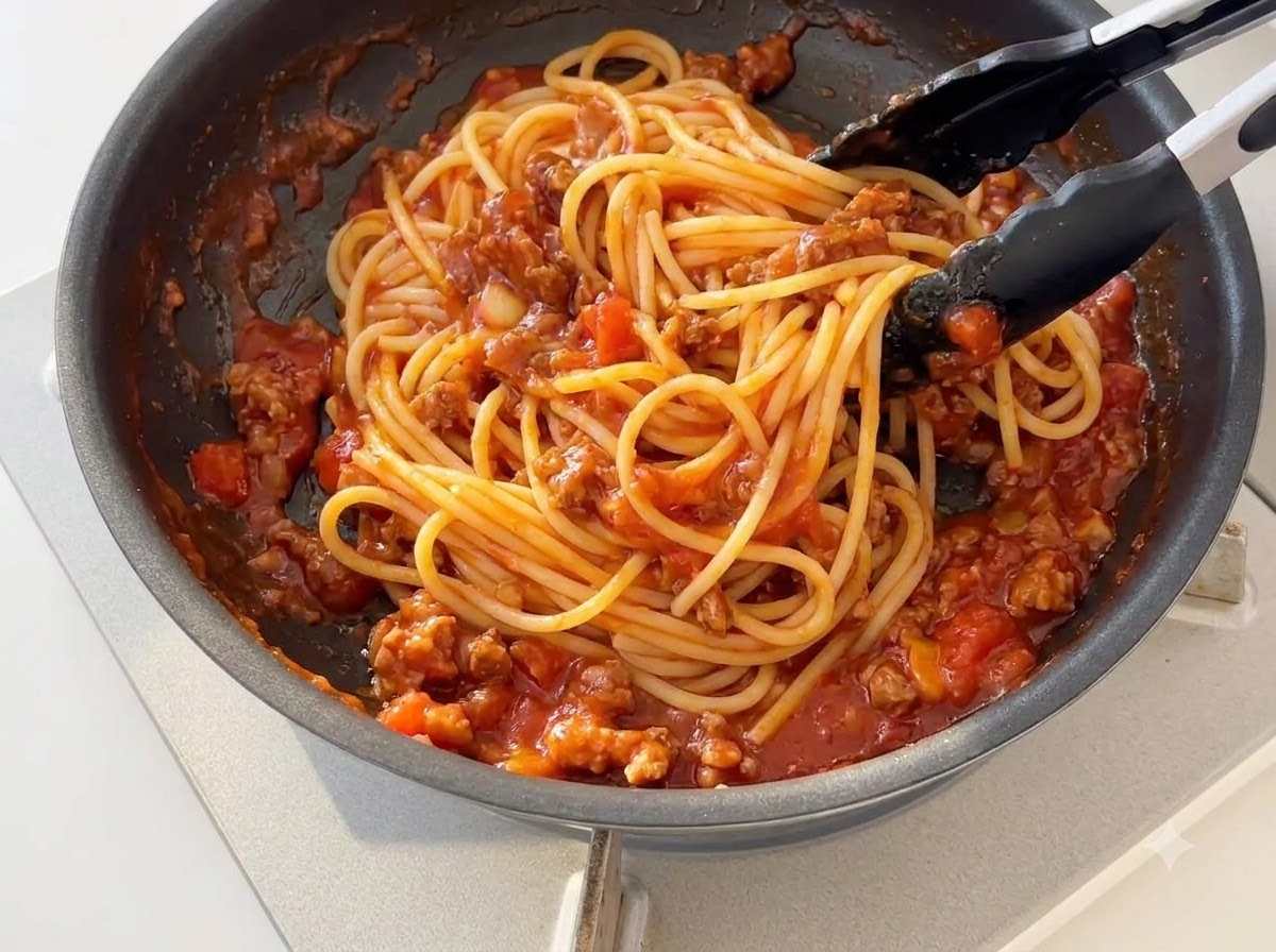 Gekochte Spaghetti-Nudeln werden mit einer schwarzen Silikonzange mit eingedickter Tomaten-Fleisch-Sauce in einer Pfanne geschwenkt.