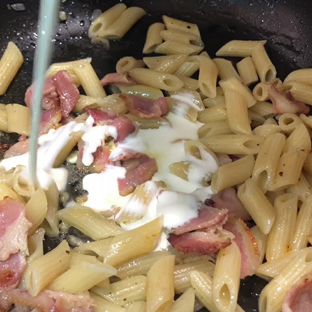 Milch und Schlagsahne werden von oben in eine Pfanne mit Penne-Nudeln und Baconstücken gegossen.