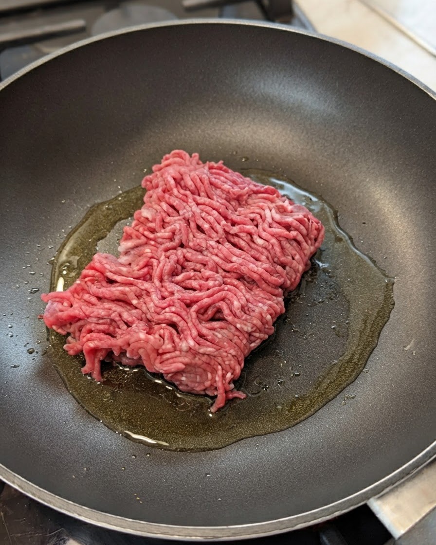 Rohes Rinderhackfleisch in einer Pfanne mit Öl, Zwiebeln und Knoblauch.