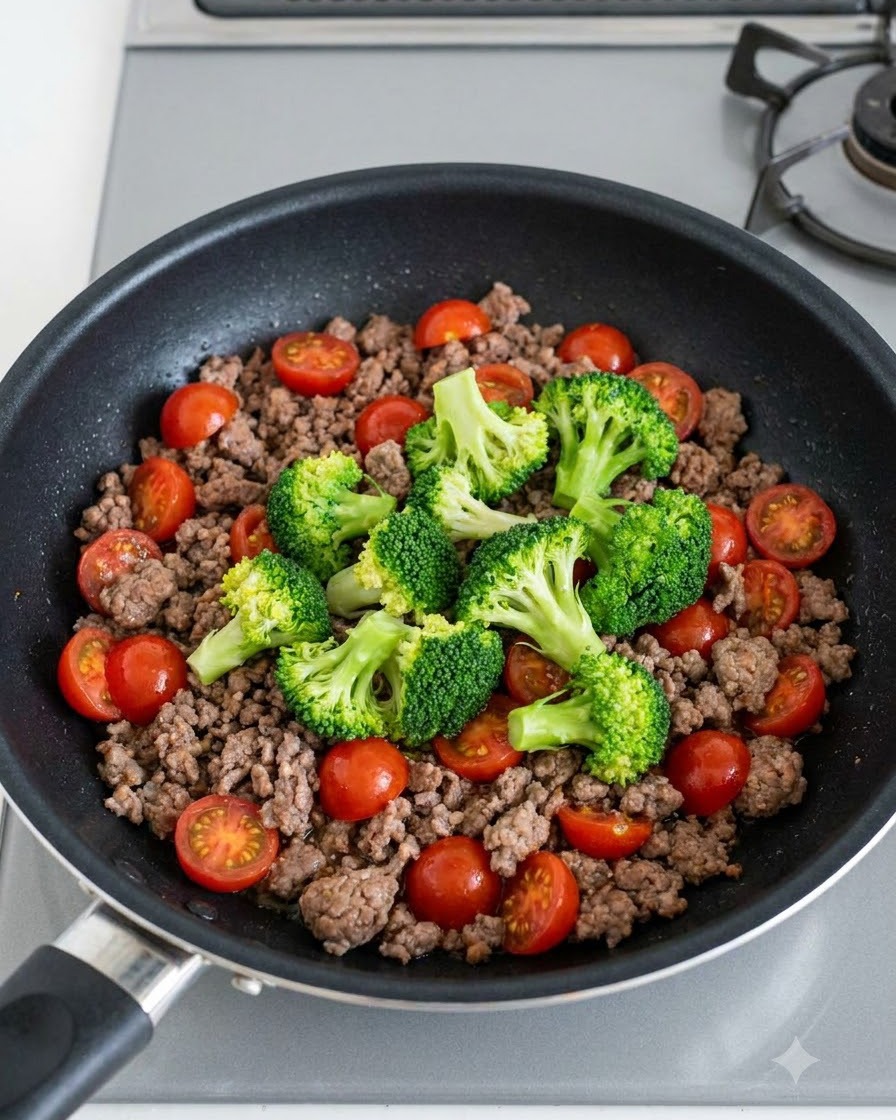 Grüne Brokkoliröschen und leuchtend rote Kirschtomaten verstreut über gebratenem Rinderhackfleisch in einer Pfanne.