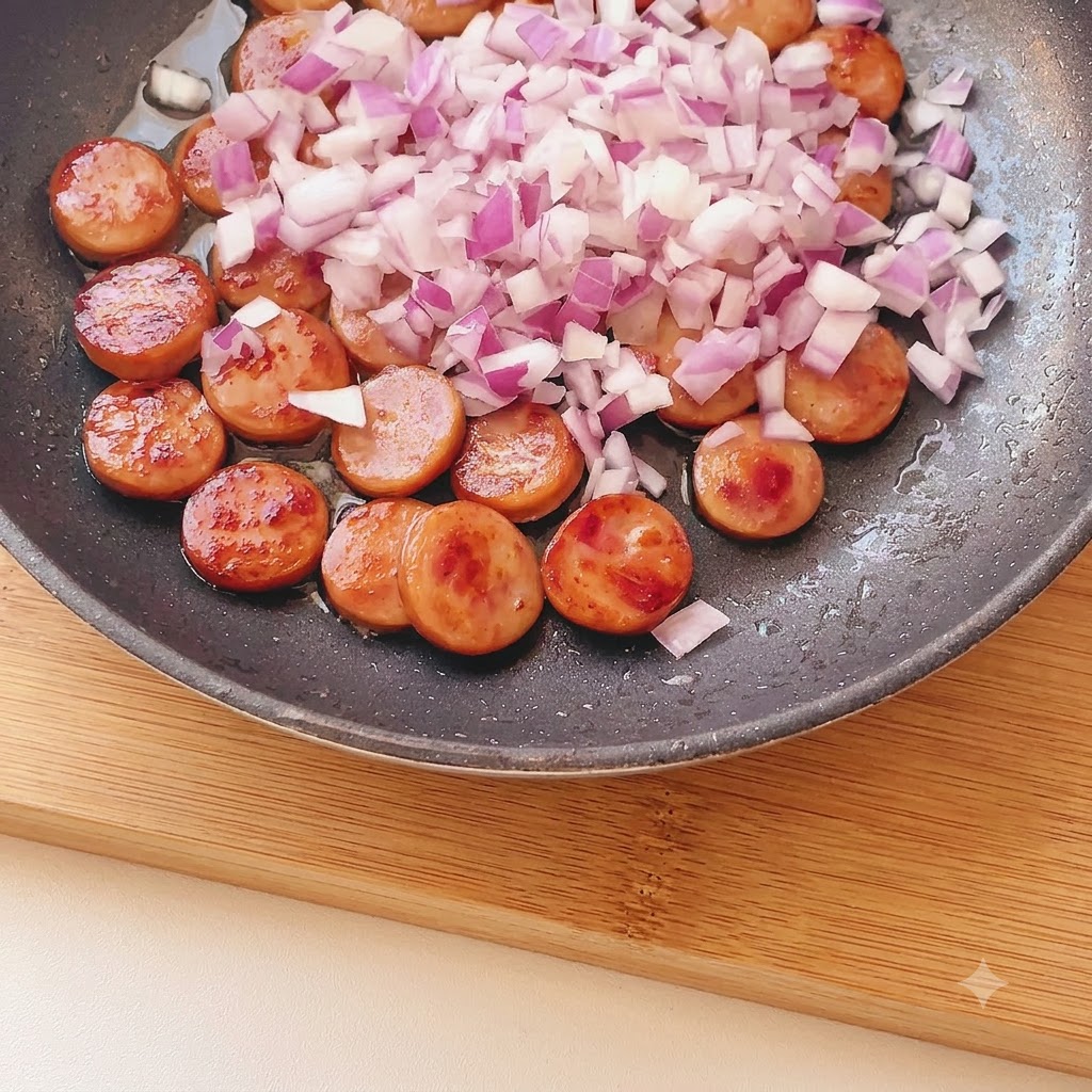 Fein gewürfelte rote Zwiebeln verteilt über gebräunten Wurststücken in einer Bratpfanne.