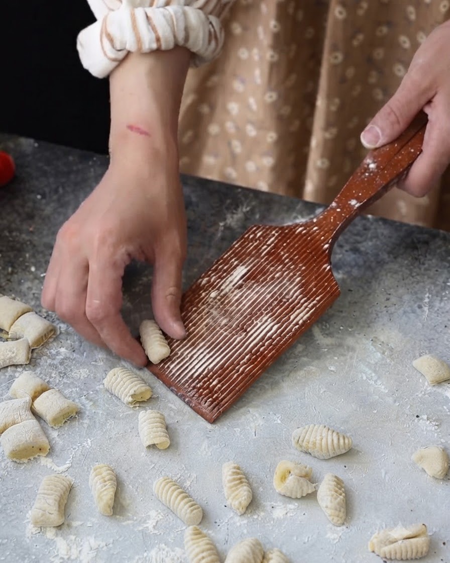 Eine Hand rollt ein kleines Teigstück über ein gerilltes Gnocchibrett aus Holz.