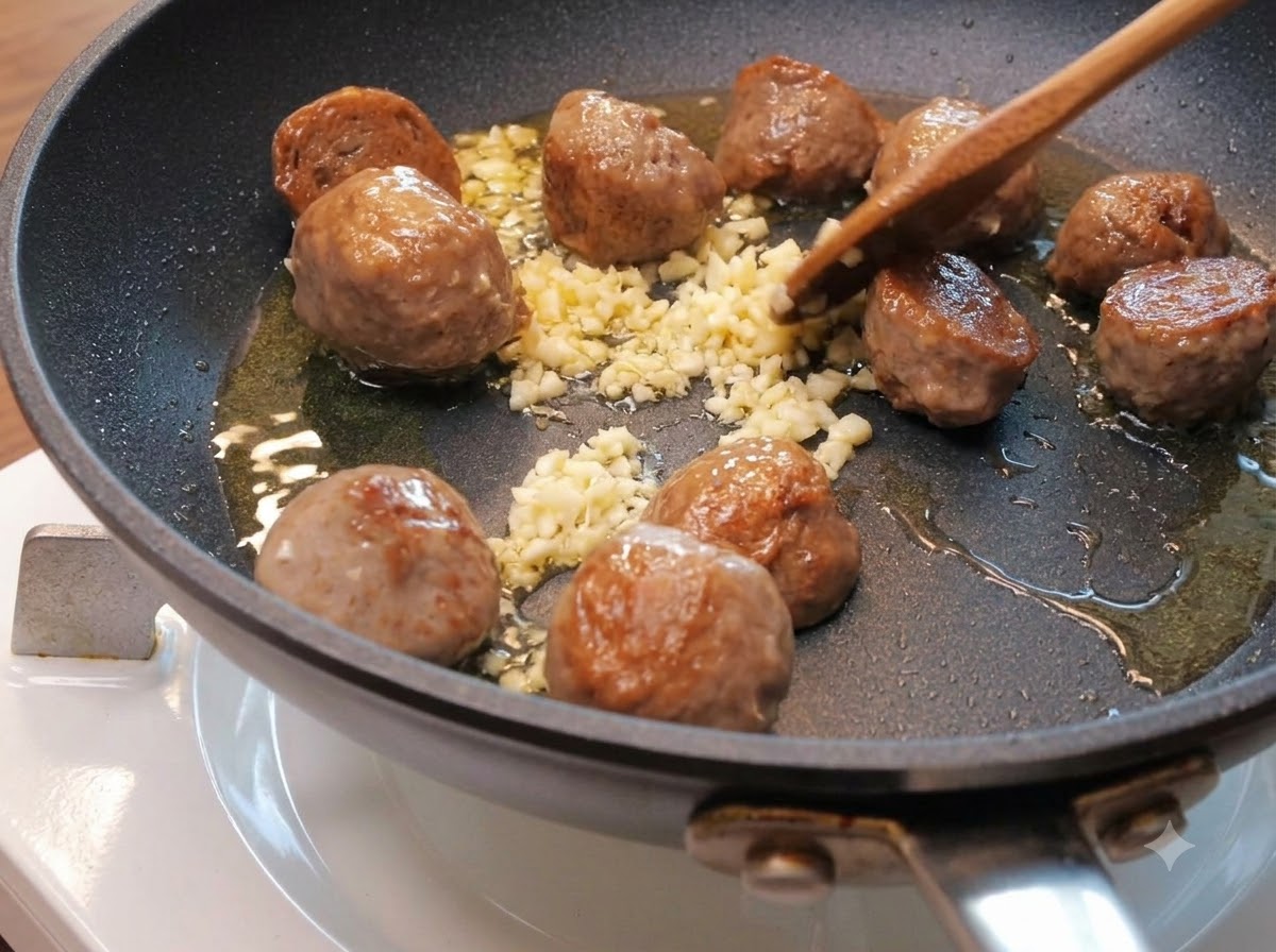 Fein gehackter Knoblauch zwischen bräunenden Rinderbällchenhälften in einer Pfanne mit Öl.