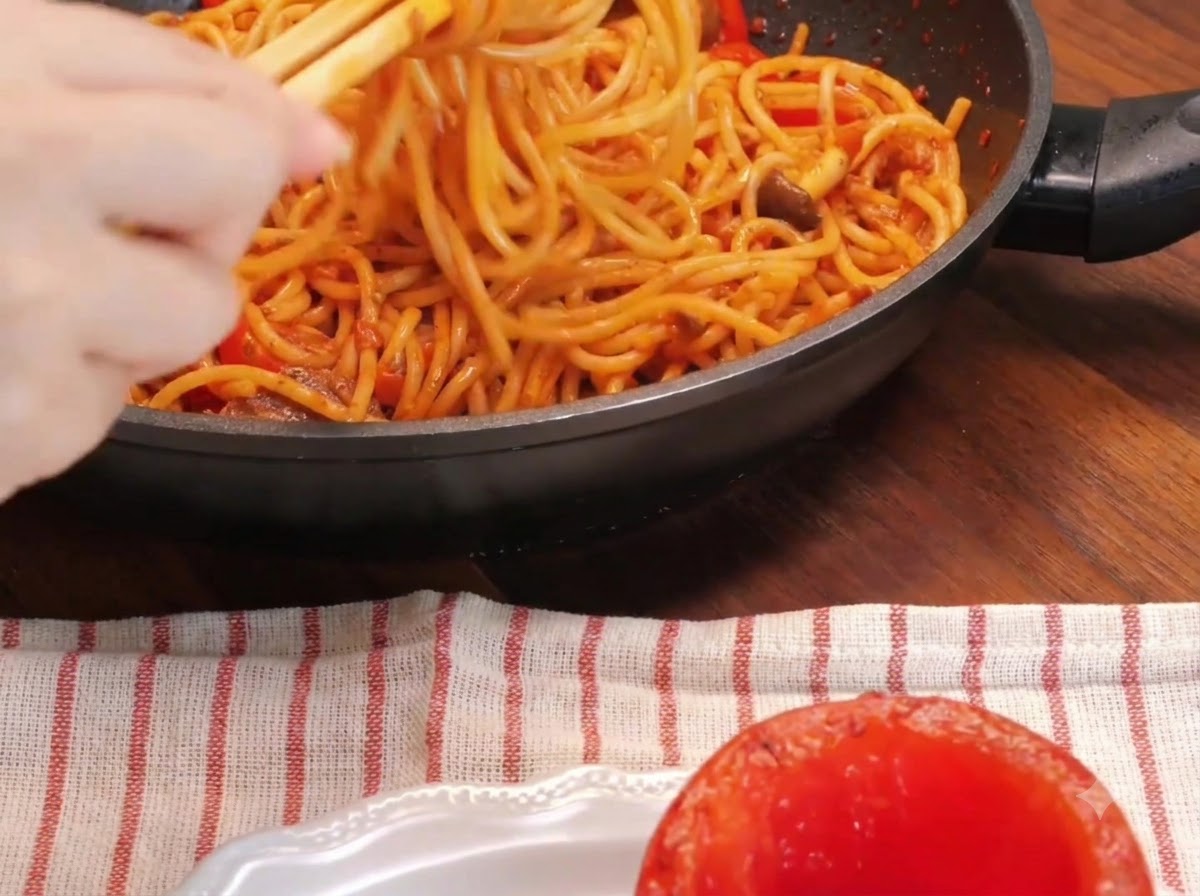 Nahaufnahme einer Hand, die Spaghetti mit Stäbchen in einer Pfanne neben einer ausgehöhlten Ofentomate auf einem Teller dreht.