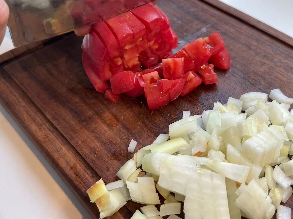 Eine frische rote Tomate wird mit einem Küchenmesser auf einem Holzschneidebrett neben gehackten Zwiebeln gewürfelt.