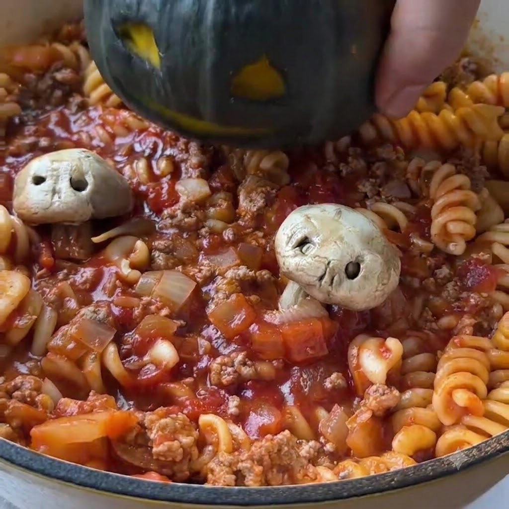 Ein ausgehöhlter, dunkelgrüner Minikürbis wird in die Mitte einer Pfanne mit gekochter Tomatenpasta und Hackfleischsauce abgesenkt.