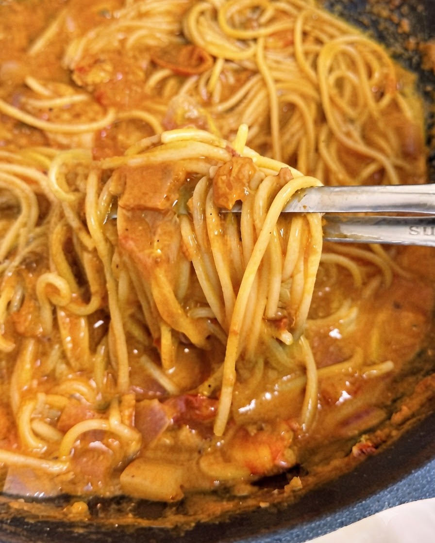 Eine Zonge hebt Spaghetti-Stränge umhüllt von cremiger rosa Tomatensauce in einem Wok an.