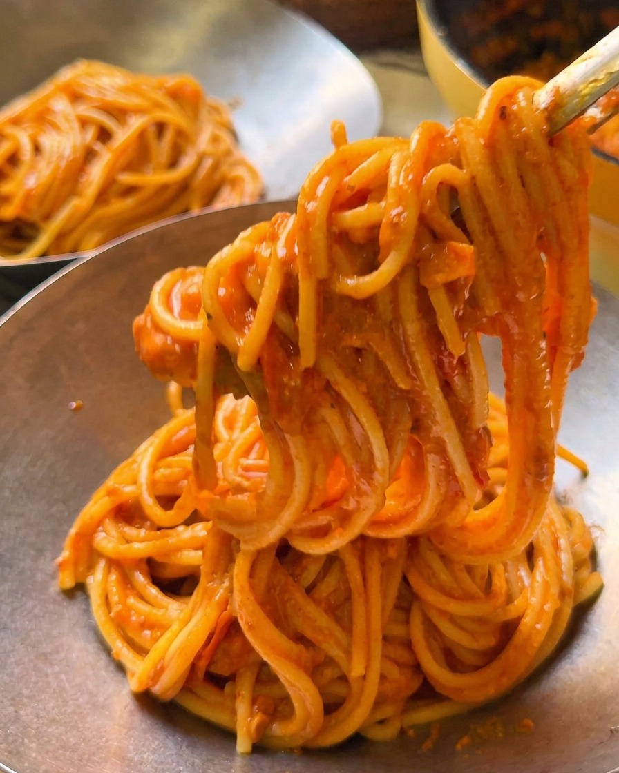 Eine Zonge hebt eine Portion cremige Tomatenspaghetti in eine polierte Servierschüssel aus Metall.