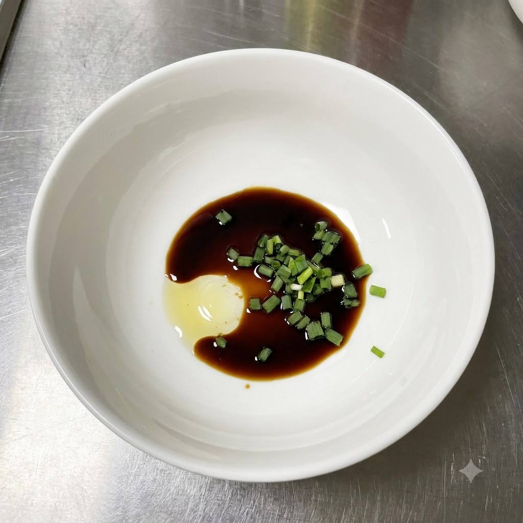 Eine weiße Schüssel mit heller Sojasoße, frisch gehackten grünen Frühlingszwiebeln und einem Klecks Würzpaste auf einer Edelstahlunterlage.