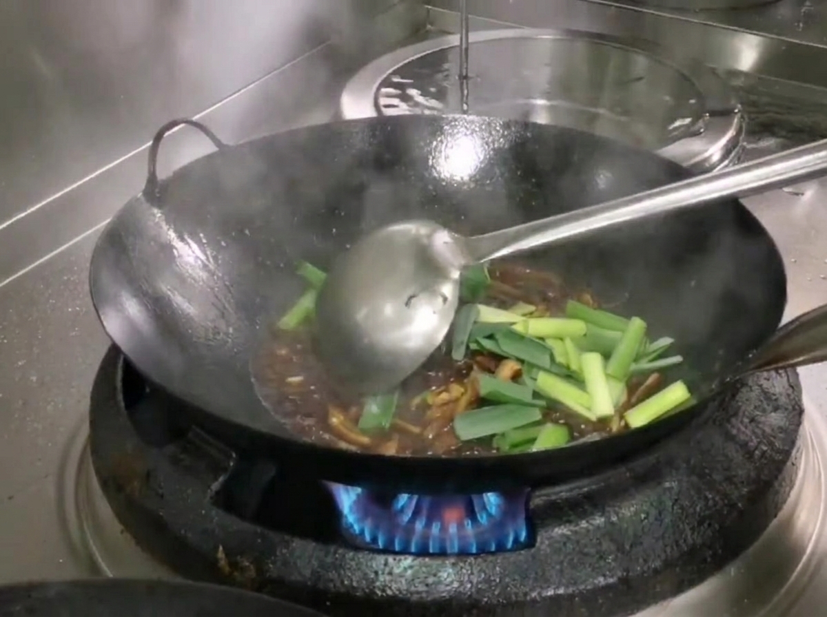 Frische grüne Abschnitte von Knoblauchtrieben werden in den Wok gegeben und mit einer Kelle unter die Zutaten gemischt.