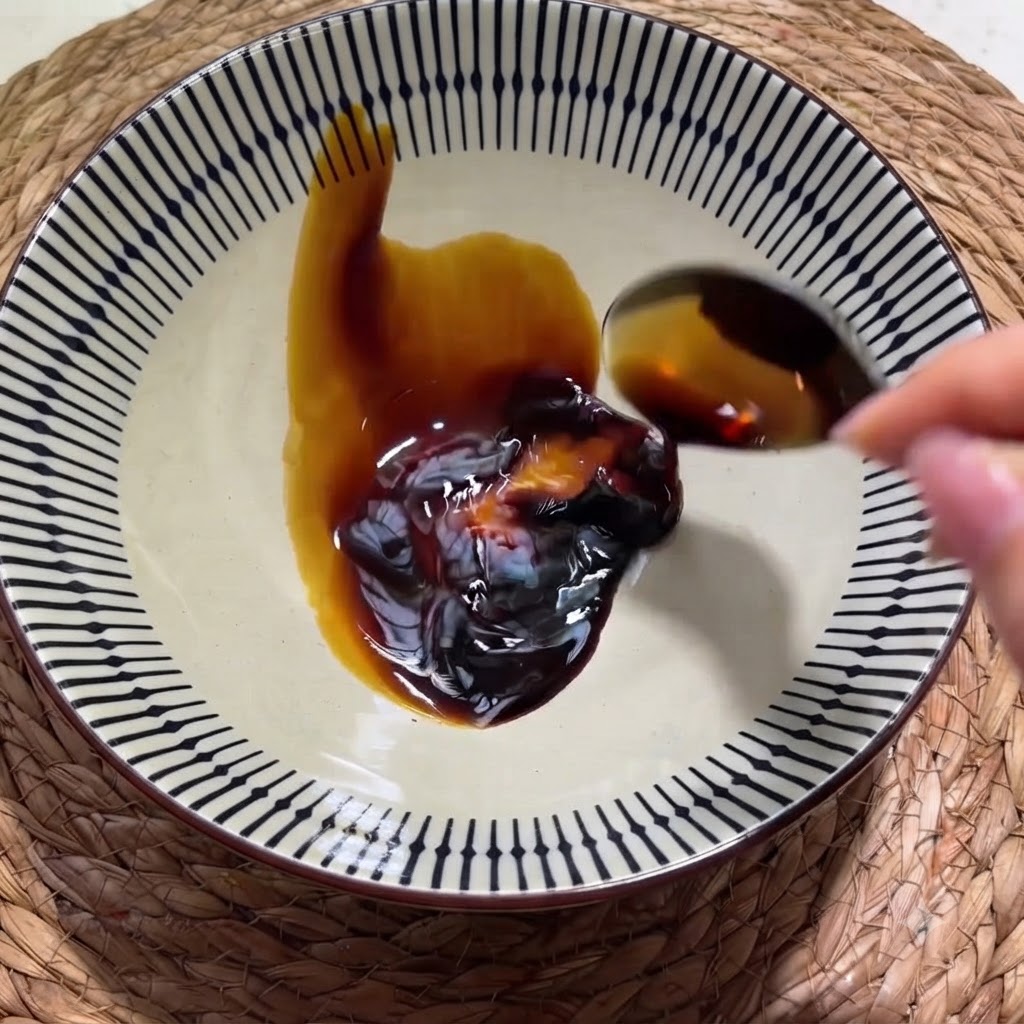 Eine Hand gießt dunkle Sojasauce von einem Löffel in eine gemusterte Keramikschüssel mit Austernsauce.