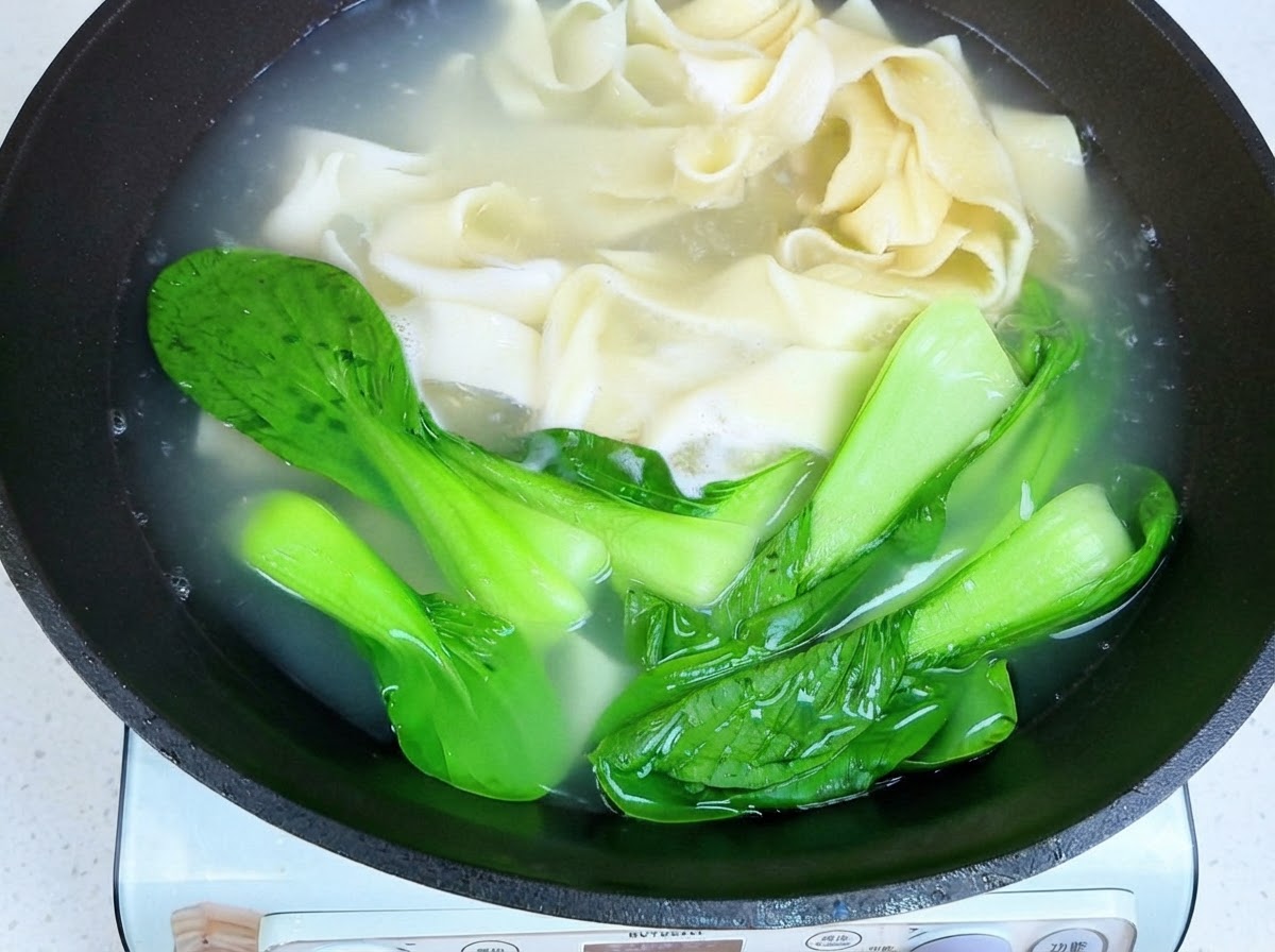 Essstäbchen heben gekochte breite Nudeln und Pak Choi aus dem heißen Wasser.