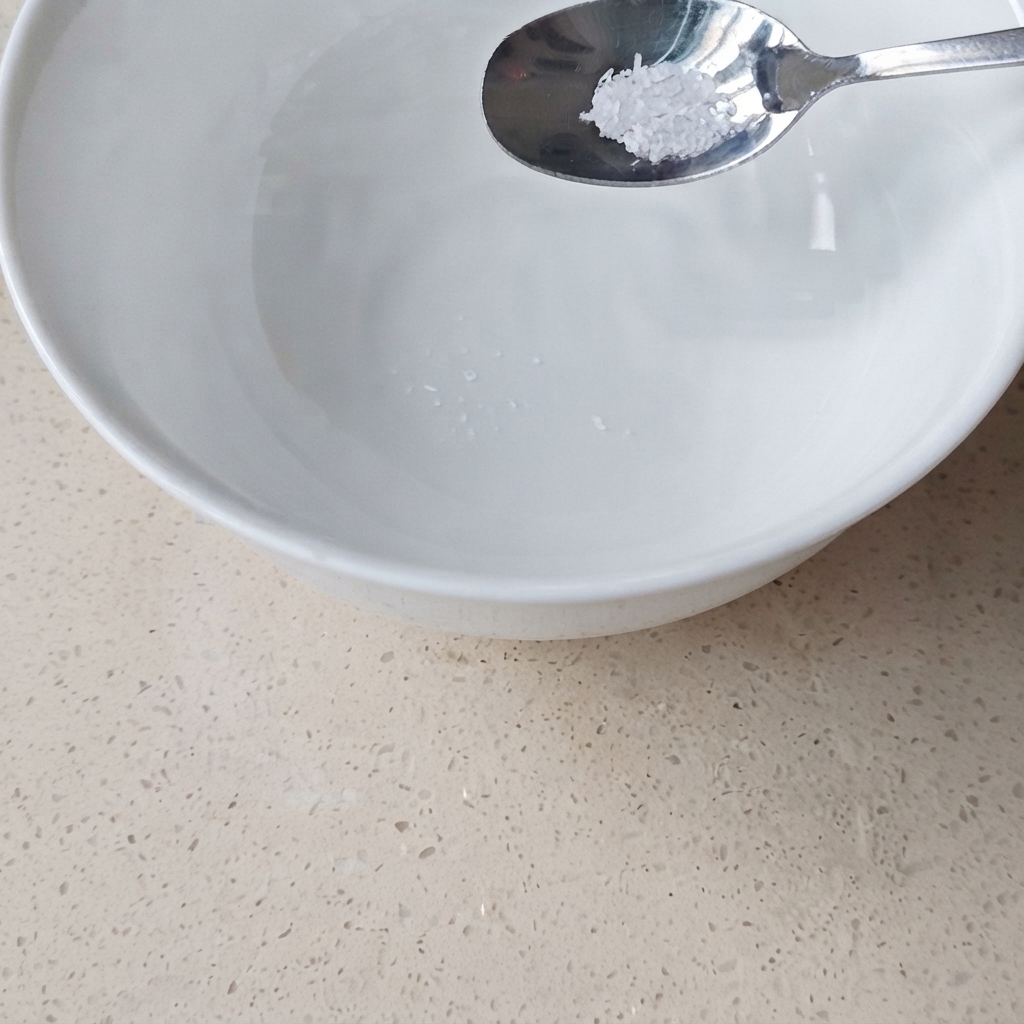 Ein Metalllöffel mit einer kleinen Menge Glutamat wird über eine weiße Keramikschüssel mit Wasser gehalten.