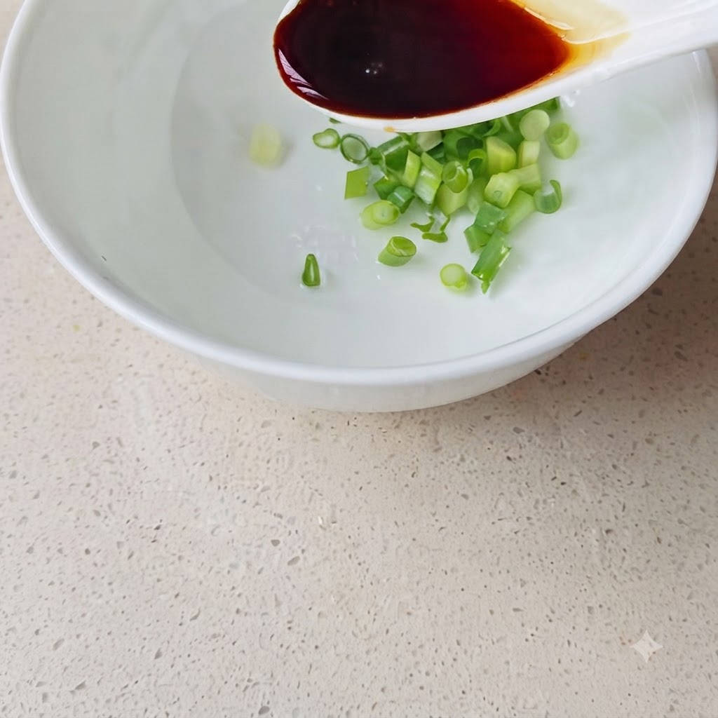 Ein weißer Löffel gießt dunkle Sojasauce in eine weiße Schüssel, die bereits gehackte Frühlingszwiebeln und Wasser enthält.