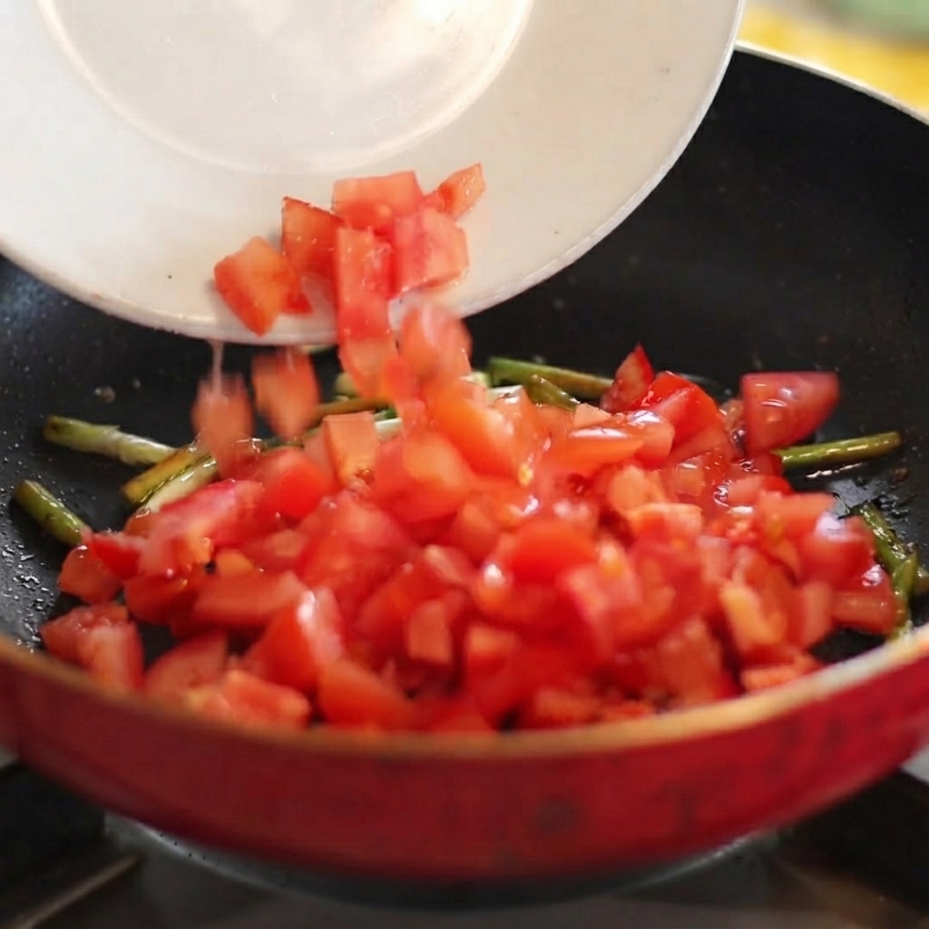 Gewürfelte rote Tomaten gleiten von einem weißen Teller in eine heiße Pfanne mit angebratenen Frühlingszwiebeln.