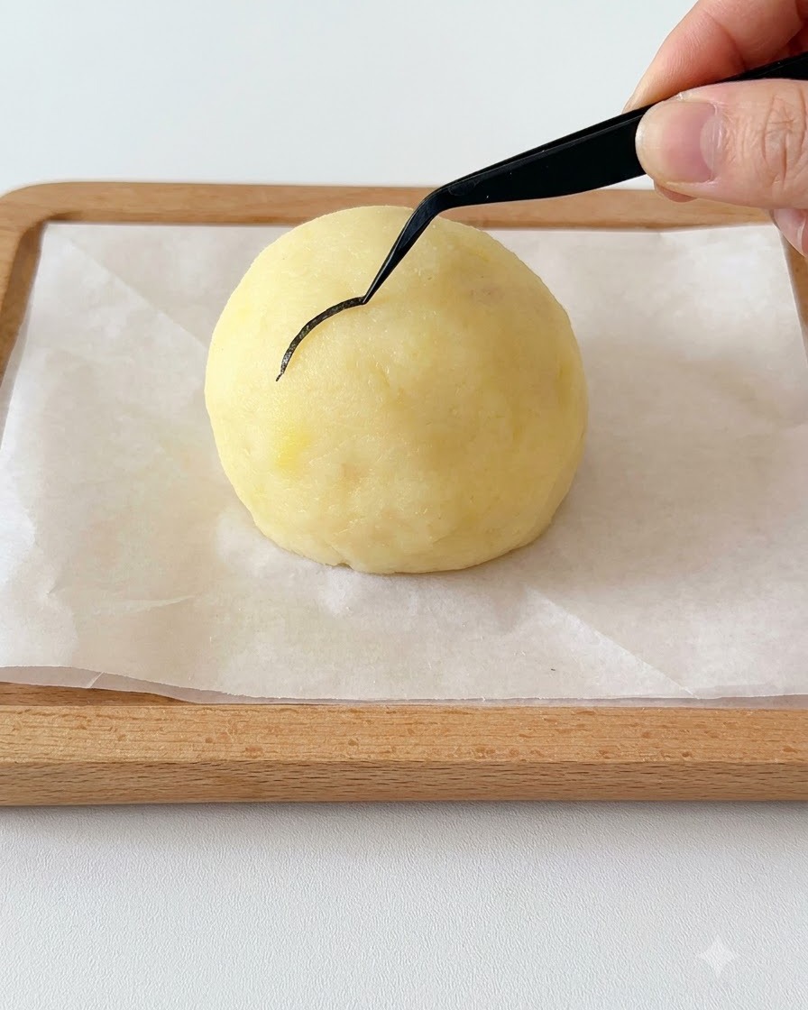 Eine Hand hält eine schwarze Pinzette und platziert ein dünnes, geschwungenes Stück Nori-Alge auf einen kugelförmigen Kartoffelkopf.