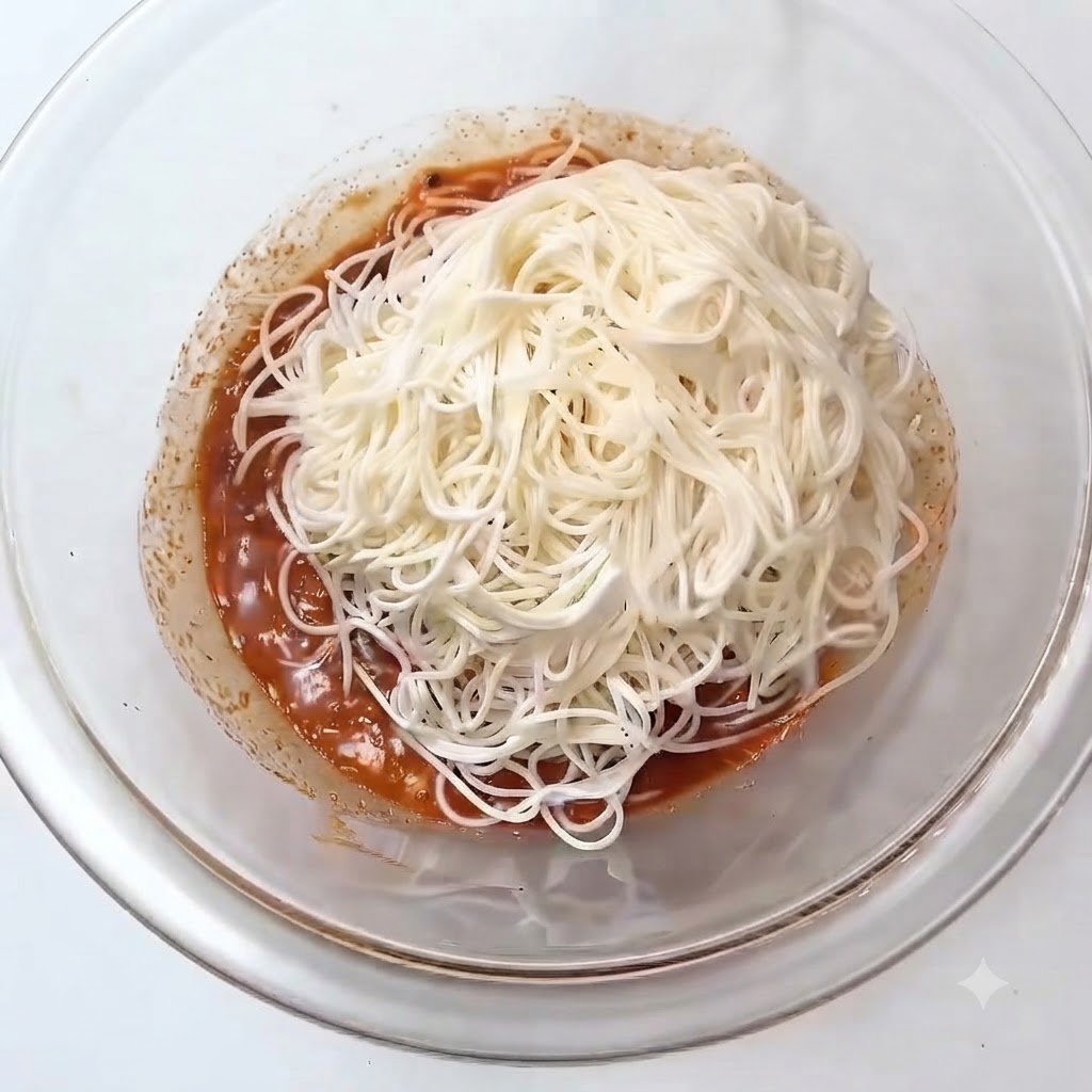 Ein Haufen weißer, gekochter Somen-Nudeln liegt auf der rötlichen Sauce in einer Glasrührschüssel.