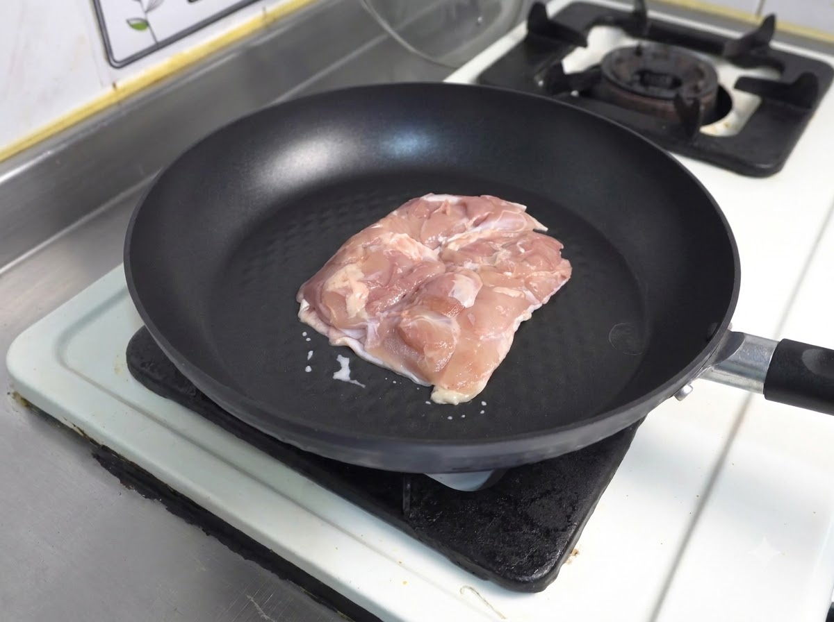 Ein Stück roher Hähnchenschenkel mit der Hautseite nach unten in einer dunklen Antihaft-Pfanne.