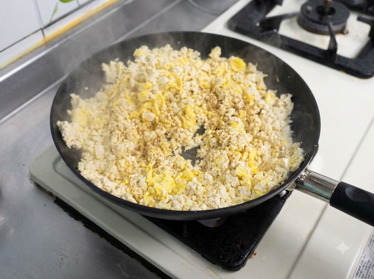 Eine Bratpfanne gefüllt mit einer Mischung aus Rührei und Tofu, die mit Gewürzen bestreut und angebraten wird.