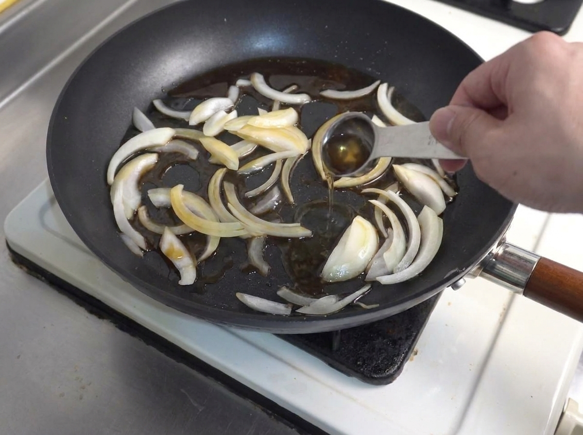 Ein Messlöffel gießt flüssige Würze in eine Bratpfanne mit angebratenen Zwiebelscheiben.