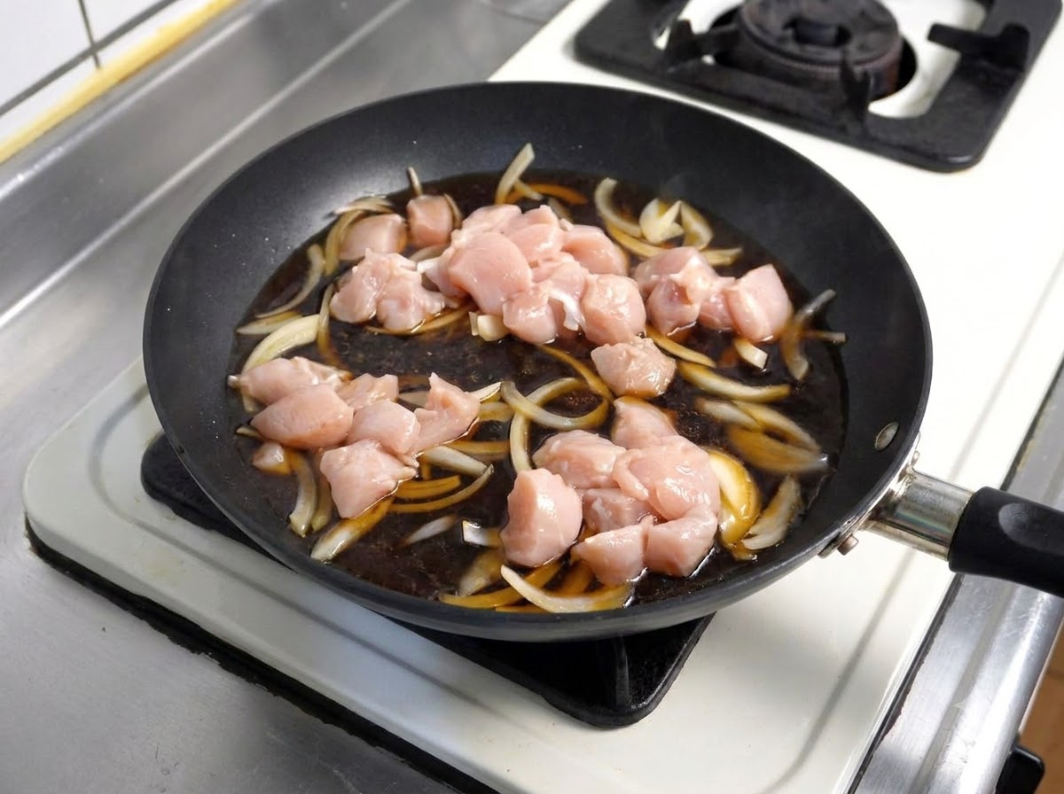 Rohe Hähnchenstücke werden in eine köchelnde dunkle Sauce mit Zwiebeln in einer Bratpfanne gegeben.