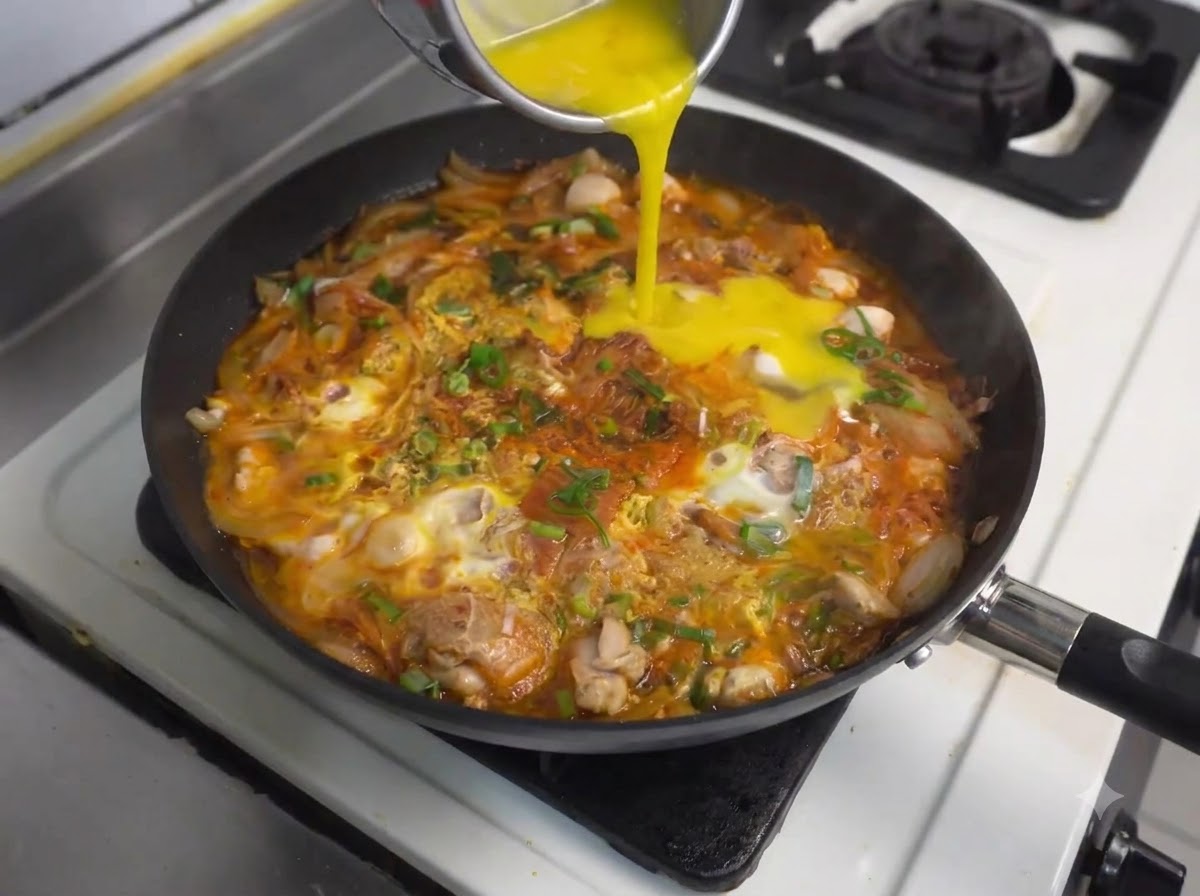 Weitere verquirlte Eier werden aus einem Behälter in die köchelnde Pfanne mit Hähnchen und Kimchi gegossen.