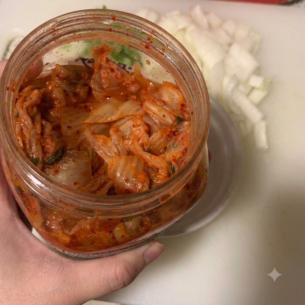 Kimchi wird mit einem Löffel in eine kleine weiße Schüssel gegeben, neben einem Behälter mit gehackten Zwiebeln.