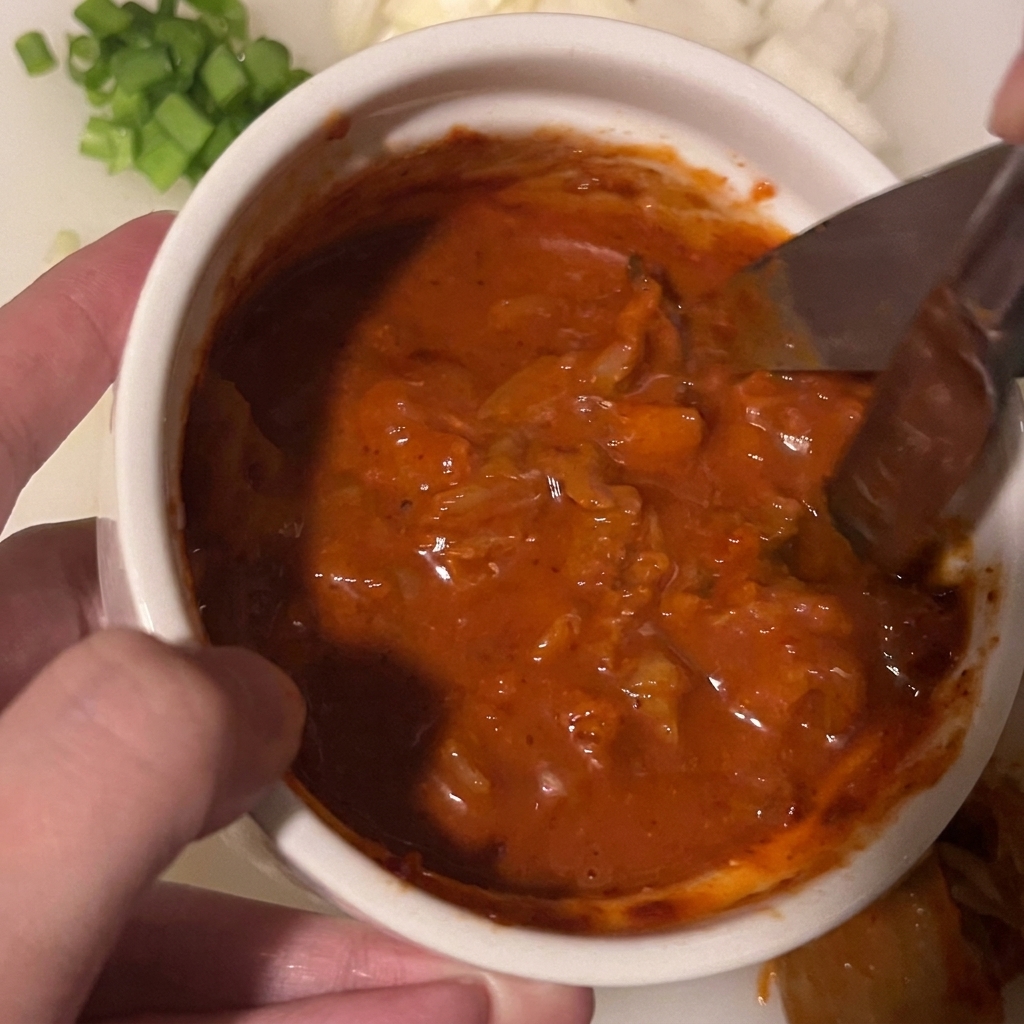 Küchenschere schneidet Kimchistücke durch, die in einer dicken roten Soßenmischung in einer kleinen weißen Schüssel liegen.