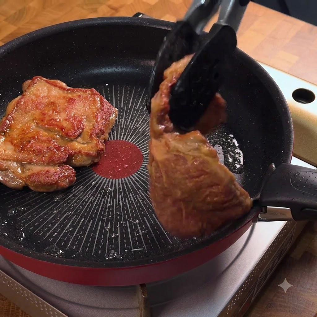 Eine Küchenzange wendet ein goldbraun gebratenes Schweinekotelett in der Pfanne.