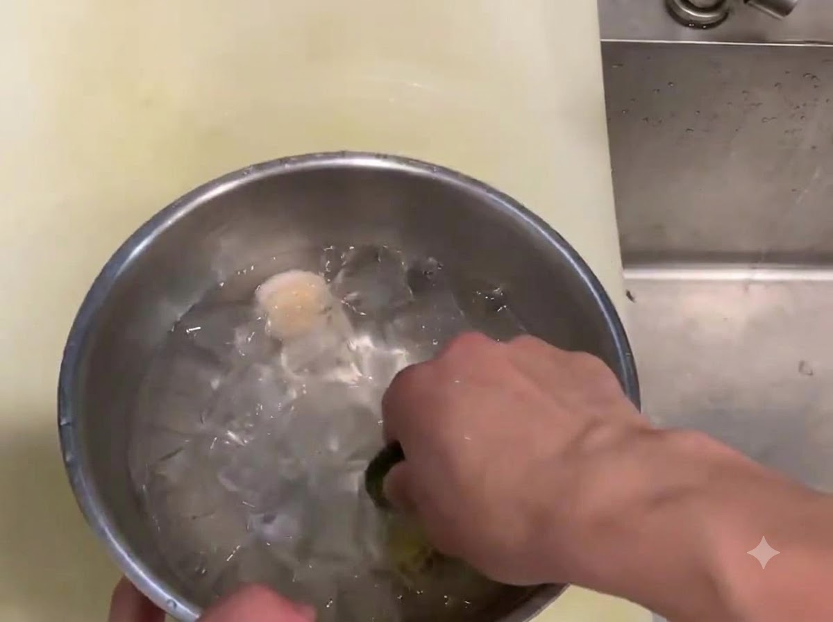 Eine Hand schwenkt ein rohes Muschelfleisch in einer Metallschüssel mit Eiswürfeln und kaltem Wasser.