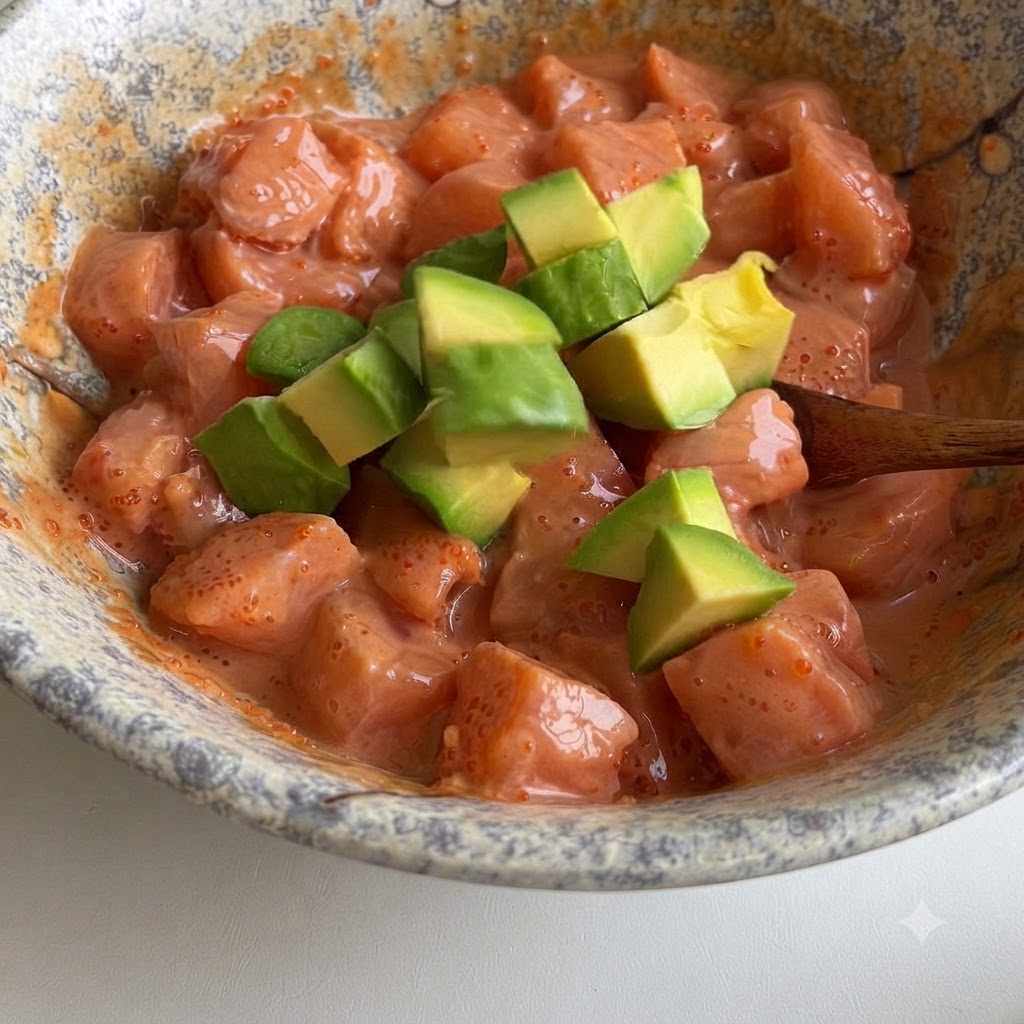 Grüne Avocadowürfel werden vorsichtig unter eine Schüssel mit Lachswürfeln in cremiger rosa Soße gehoben.