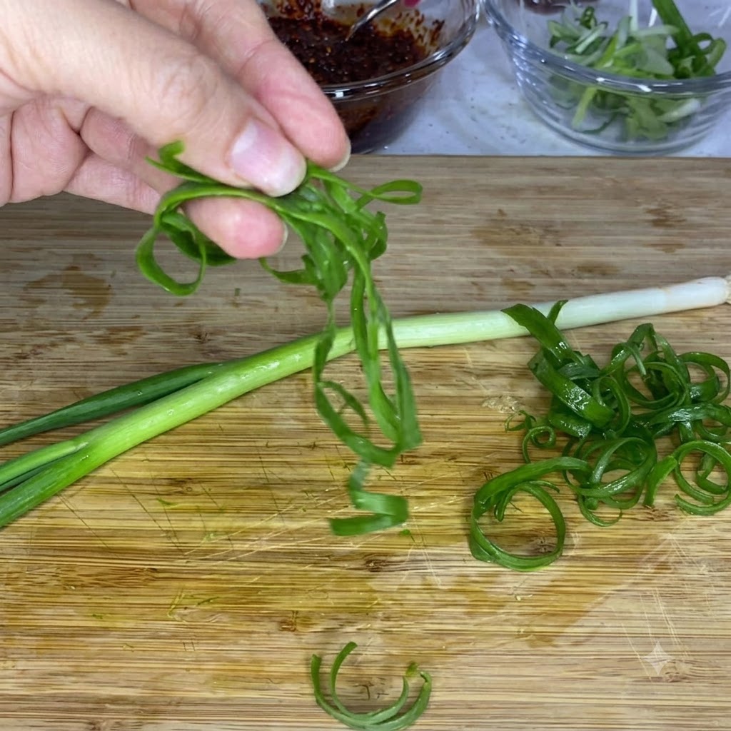 Eine Hand hält gekräuselte grüne Frühlingszwiebelstreifen über ein Holzschneidebrett, mit Glasschüsseln mit Soße und geschnittenen Zwiebeln im Hintergrund.