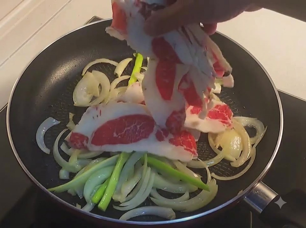 Eine Hand legt dünne Scheiben rohen Rinderbauchs in eine Bratpfanne mit geschnittenen Zwiebeln und Frühlingszwiebeln.