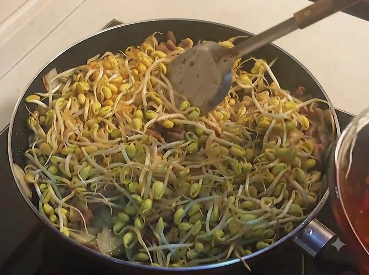 Ein Schöpflöffel gibt dunkelrote, scharfe Sauce über eine volle Pfanne mit gelben Sojasprossen.