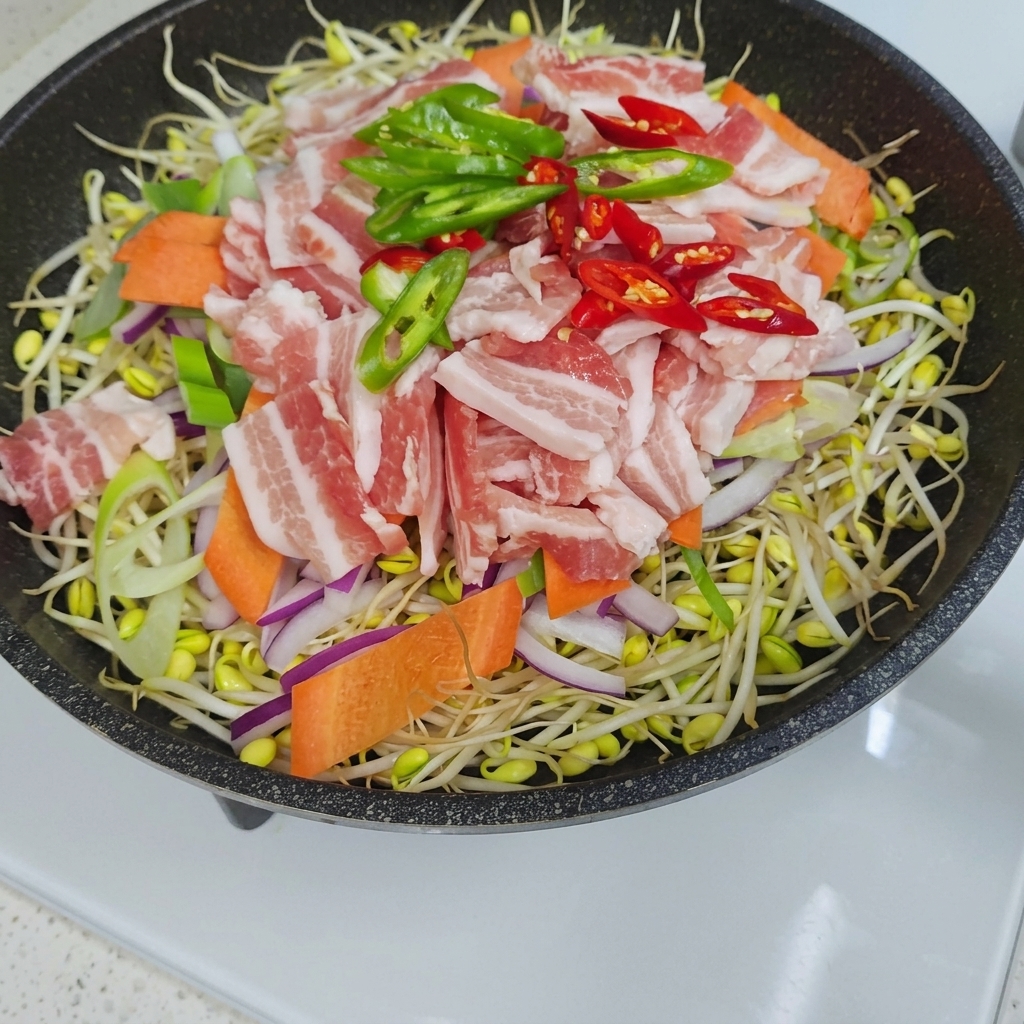 Eine Ansicht von oben auf eine Pfanne gefüllt mit Bohnensprossen, bedeckt mit Zwiebeln, Karotten, Schweinebauchstreifen sowie grünen und roten Chilistücken.
