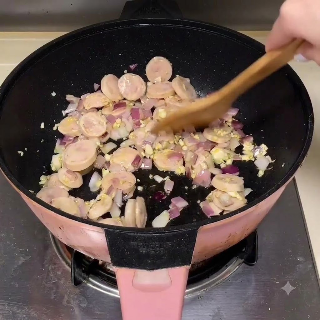 Sausage slices being mixed with onions and garlic in a pan using a wooden spatula.