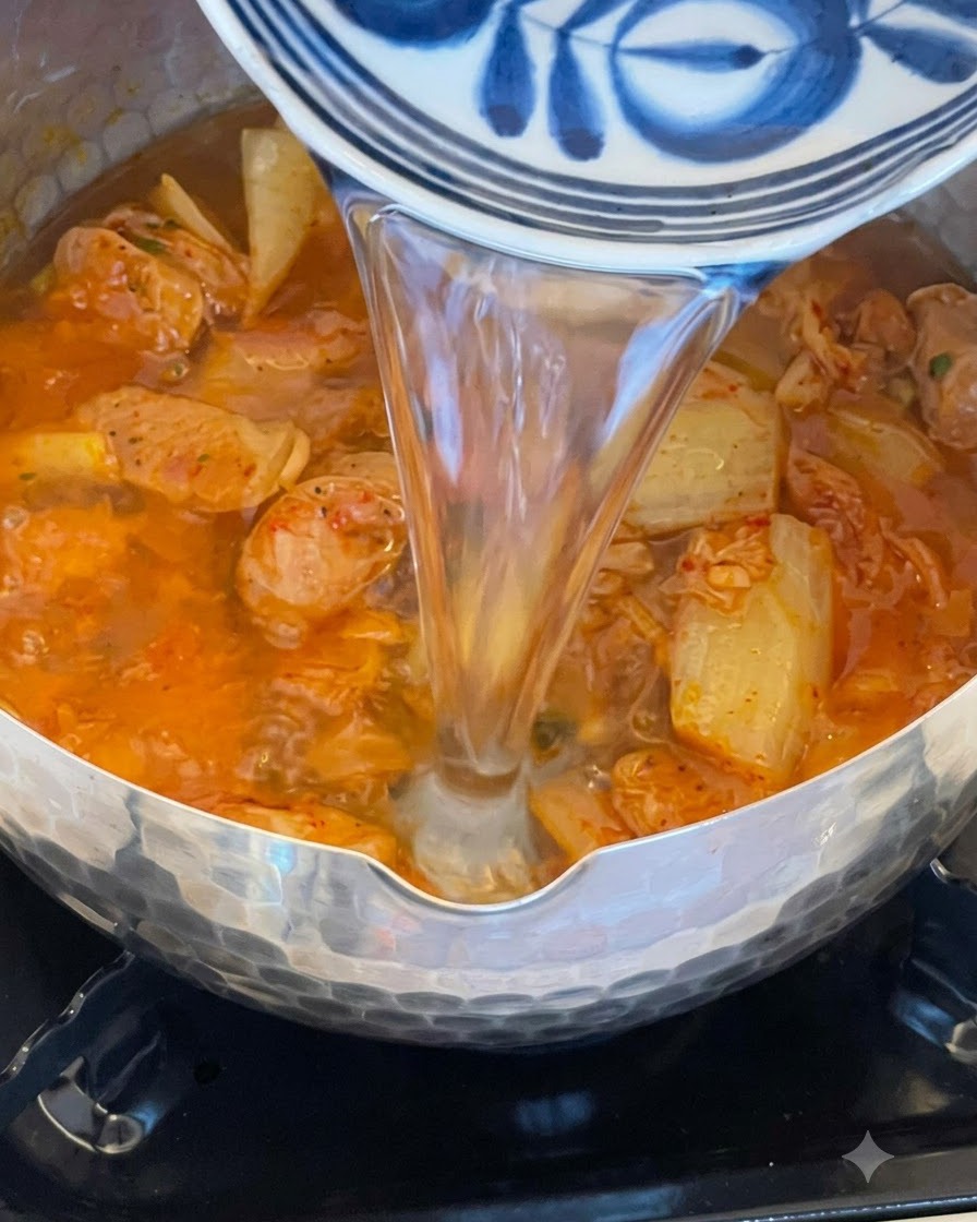 Wasser gießt aus einer dekorativen blau-weißen Schüssel in einen Topf gefüllt mit blubbernder roter Kimchi-Wurst-Brühe.