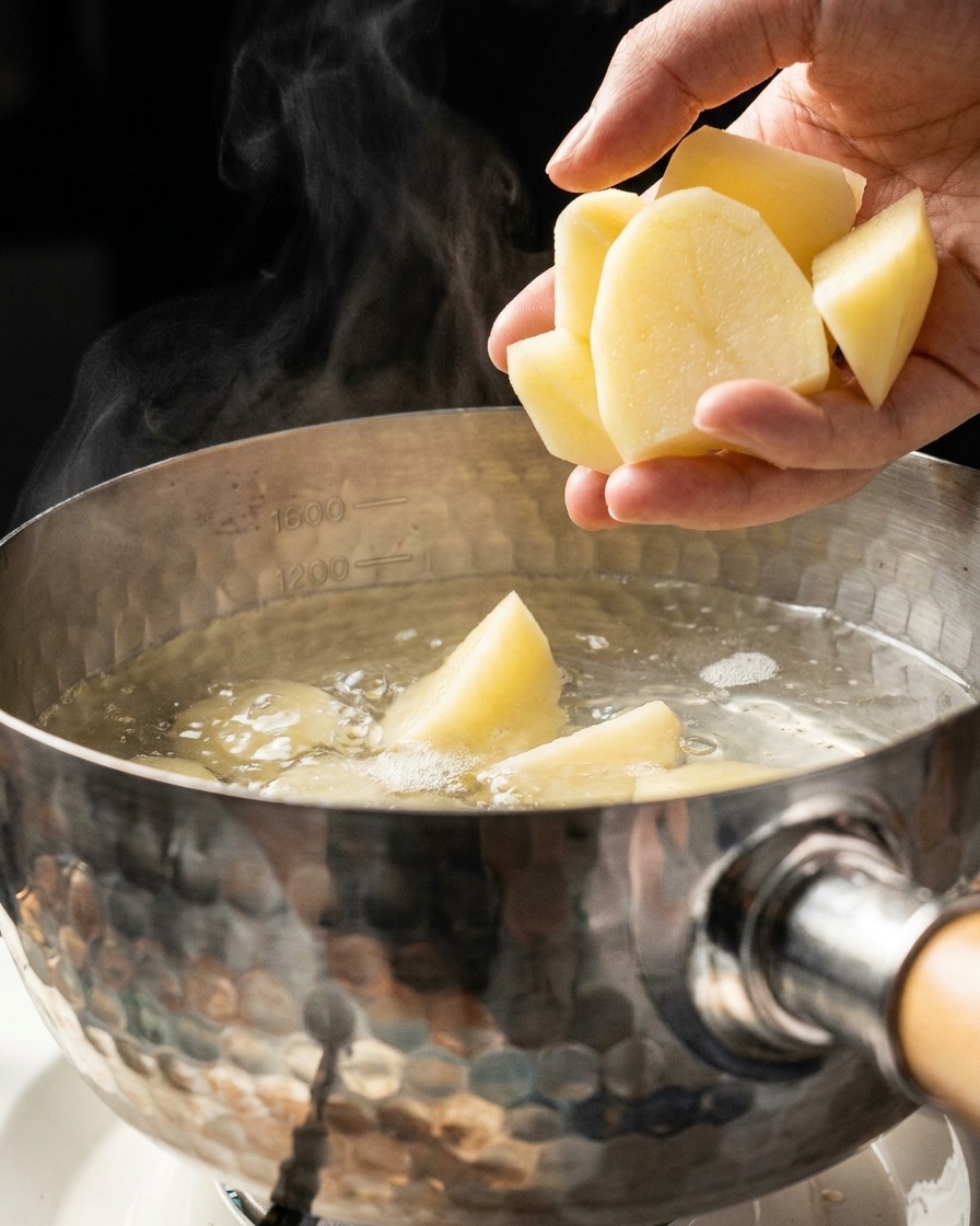 Eine Hand wirft geschnittene Kartoffelstücke in einen mit kochendem Wasser gefüllten Metalltopf, aus dem Dampf aufsteigt.