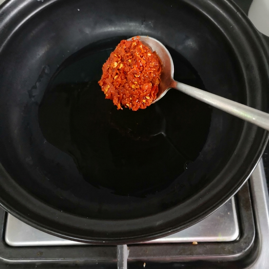 Ein Löffel rote Chilipaste wird in einen schwarzen Tontopf auf dem Herd gegeben.
