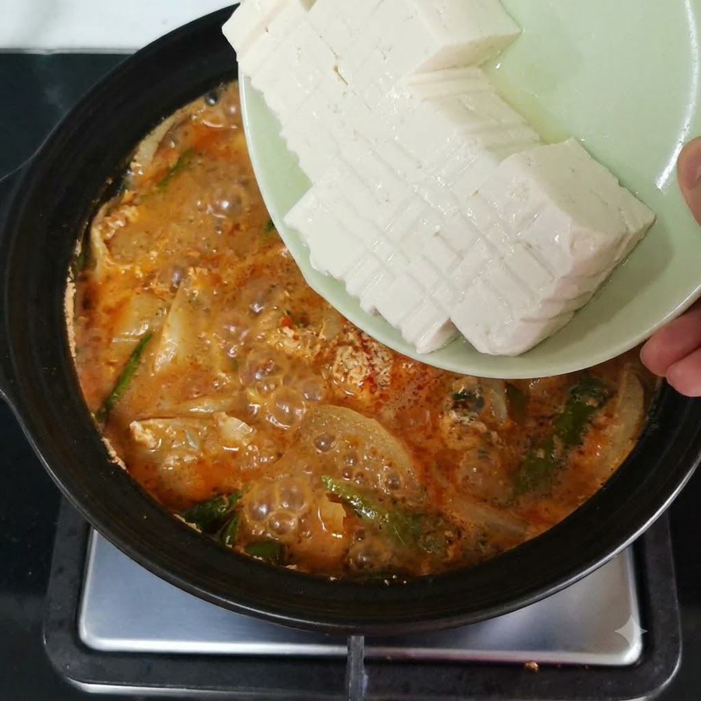 Geschnittener Seidentofu wird von einem Teller in den blubbernden Topf mit der orangeroten Brühe und dem Gemüse gekippt.