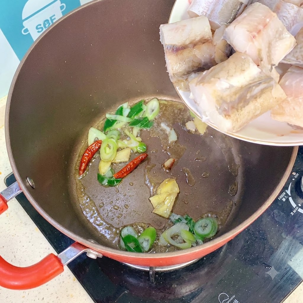 Seelachsstücke werden von einem weißen Teller in einen Topf mit angebratenen Frühlingszwiebeln, Ingwer und getrockneten Chilis gegeben.
