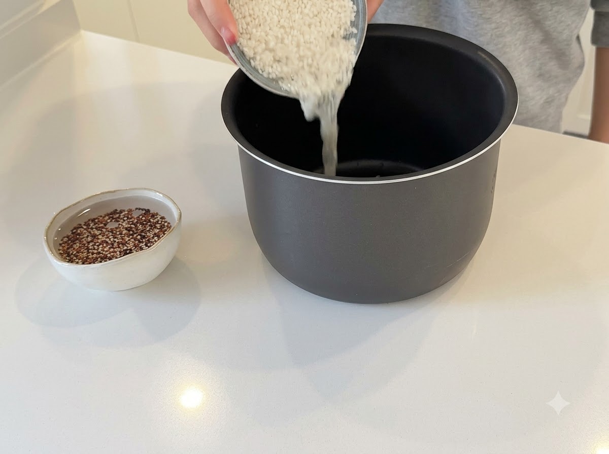 Hände füllen weißen Reis in den dunklen Topf eines Reiskochers, daneben weicht Quinoa in einer kleinen Schale ein.