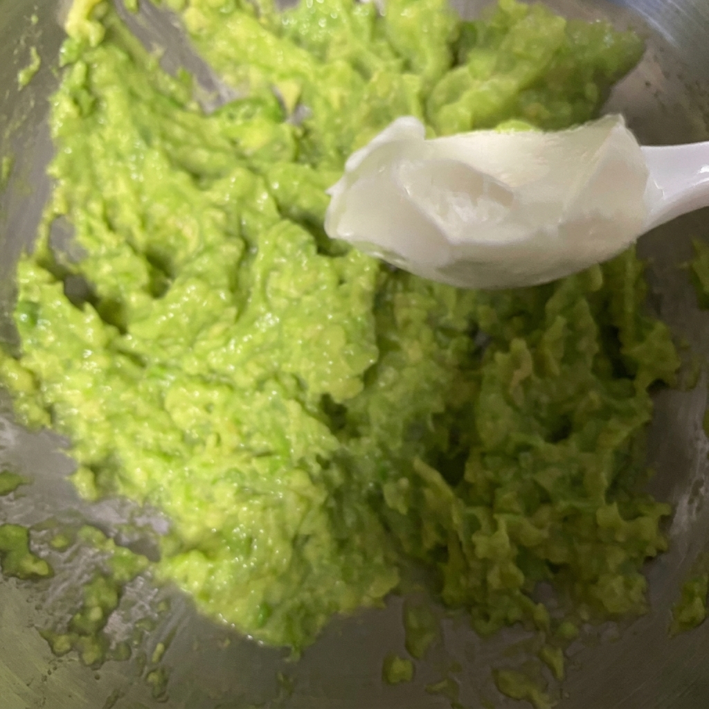 Ein weißer Löffel schöpft dicken griechischen Joghurt und gibt ihn in eine Schüssel mit frisch zerdrückter Avocado.