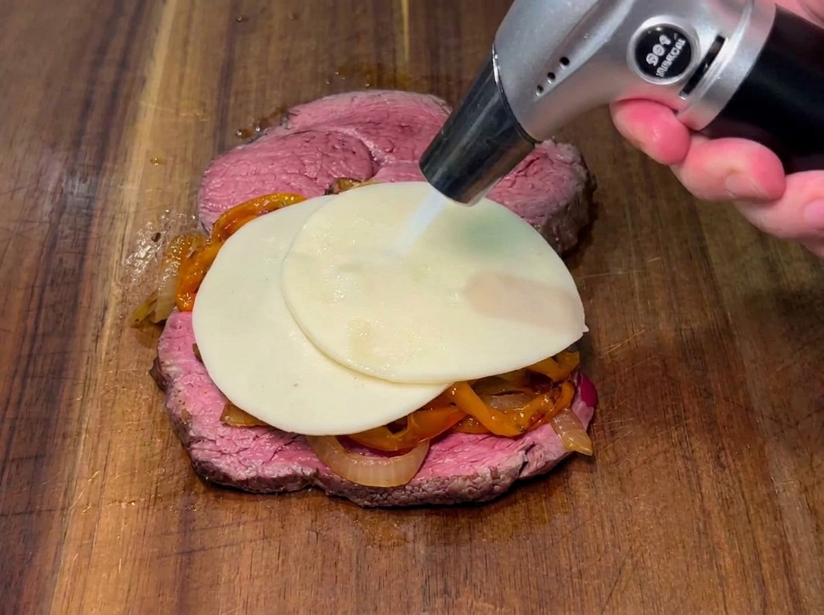 Eine Scheibe Käse wird mit einem Bunsenbrenner über Paprika und Zwiebeln auf einem Steak geschmolzen.