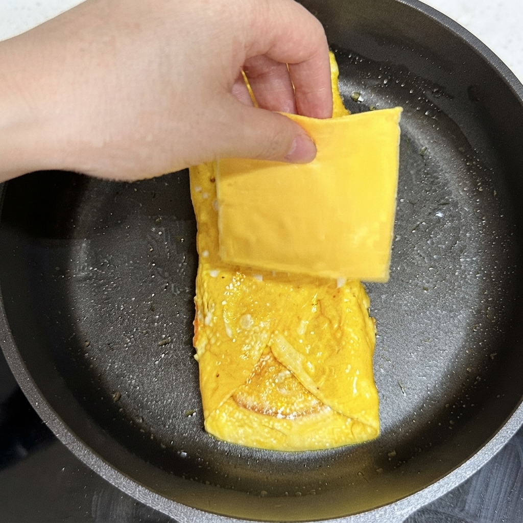Eine Hand legt eine quadratische Scheibe gelben Käse auf das gebratene Eierpaket in der Bratpfanne.
